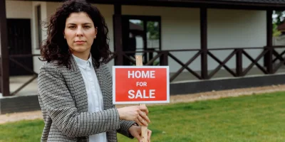 Femme tenant une pancarte "Home for Sale" devant une maison — frais de vente maison
