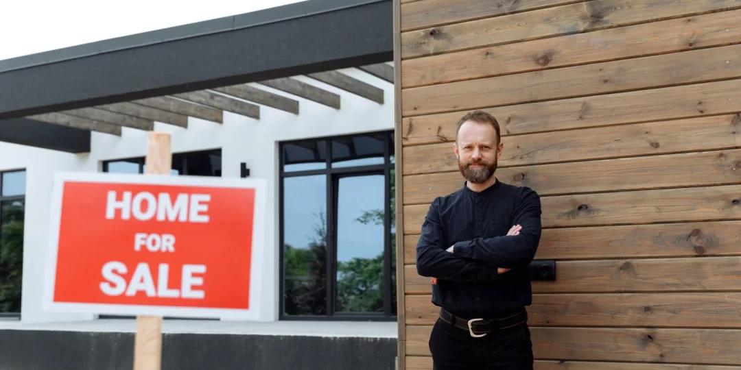 Homme souriant devant une maison avec un panneau "À vendre" — taux-immobilier